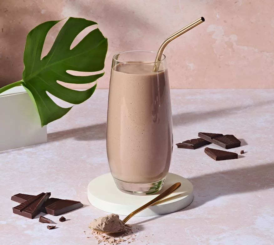 Tall glass of chocolate smoothie with a metal straw on a white circular coaster, surrounded by broken dark chocolate and a scoop of cocoa powder.