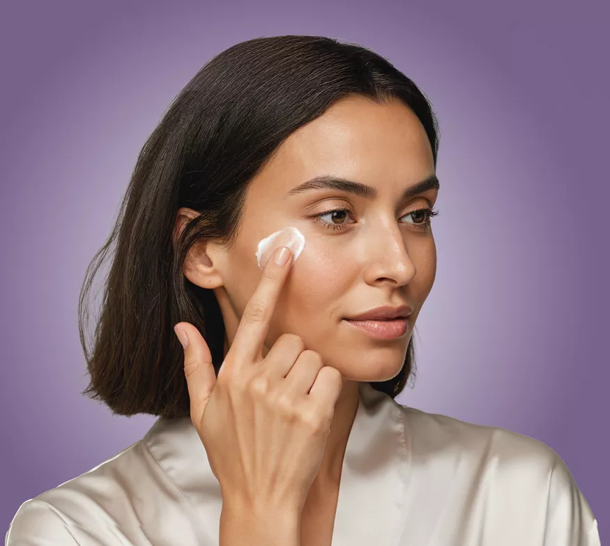 Woman with short dark hair applying cream to her cheek with a cotton pad against a purple background (skincare routine).